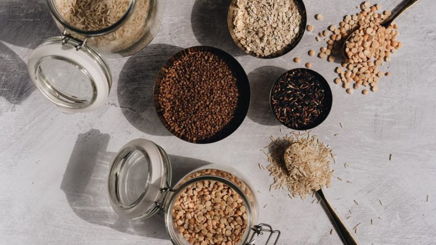 A top view of assorted grains and pulses in jars and bowls on a textured surface with shadow play.